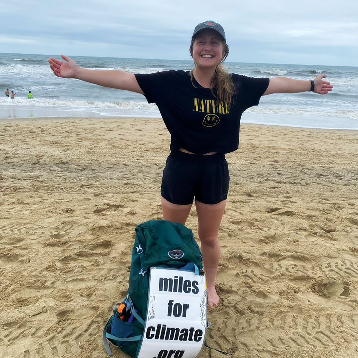 a smiling girl wearing a nature shirt holds her arms out wide in front of the oean. Her backpack reads "miles for climate .org"
