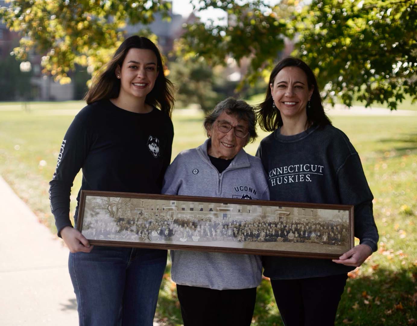 Hanna Basile, Barbara Graf Larson, and Roberta Larson Basile pose for UConn’s 2021 public service TV ad with a photo of the entire class of 1921 at what was then Connecticut Agricultural College.