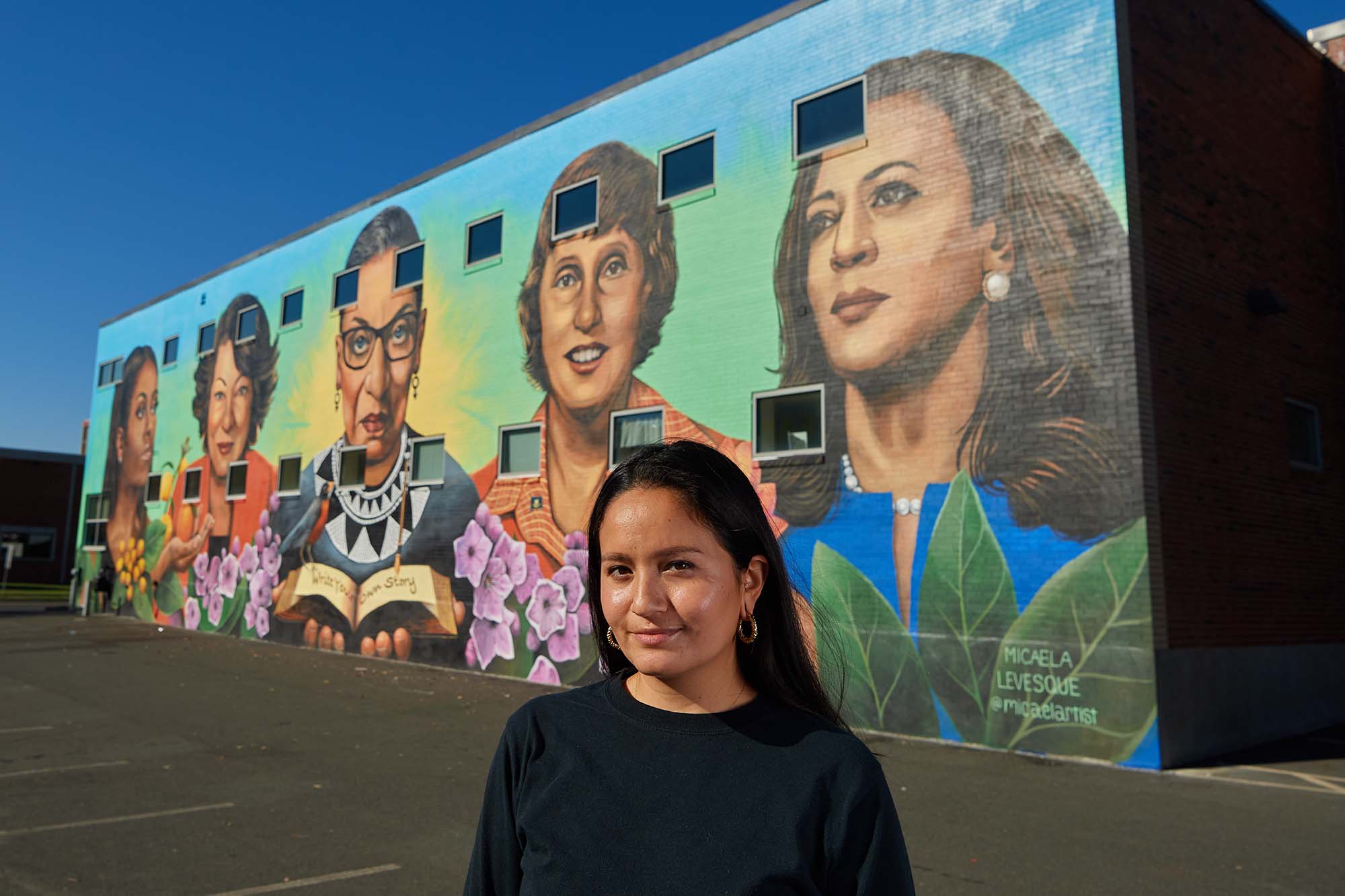 Micaela Levesque stands in front of her mural of painted historical women