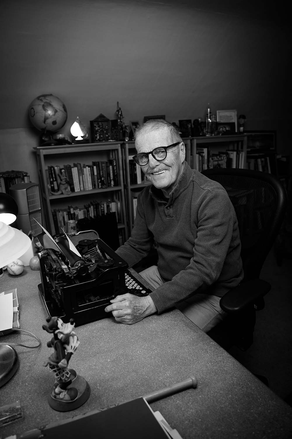 A black-and-white photo of Leigh Montville ’65 (CLAS). He sits at a desk in front of a typewriter, smiling.