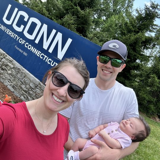 Melissa, Justin, and baby Hannah Worth, pose for a photo in front of UConn signage