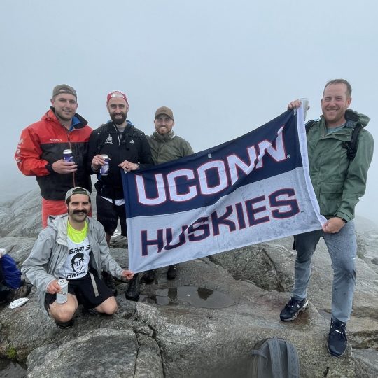 Mike Jacovino and Chris Cocozza, John Moore, Harrison Holtz and Mike Giunta stand together and hold onto a UConn Huskies flag
