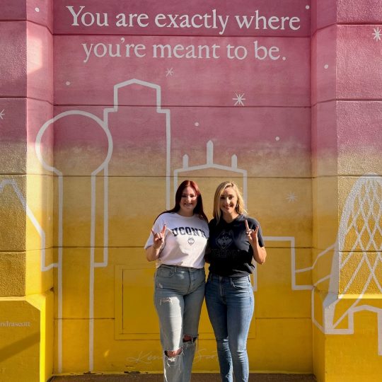 Julia Carter and Anna (Neupert) Wildfeuer stand in front of Dallas mural that states, "You are exactly where you're meant to be."