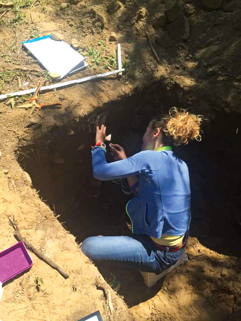 Matarazzo sits insidr carved opening in the ground surrounded by research paraphernalia while collecting archeological samples