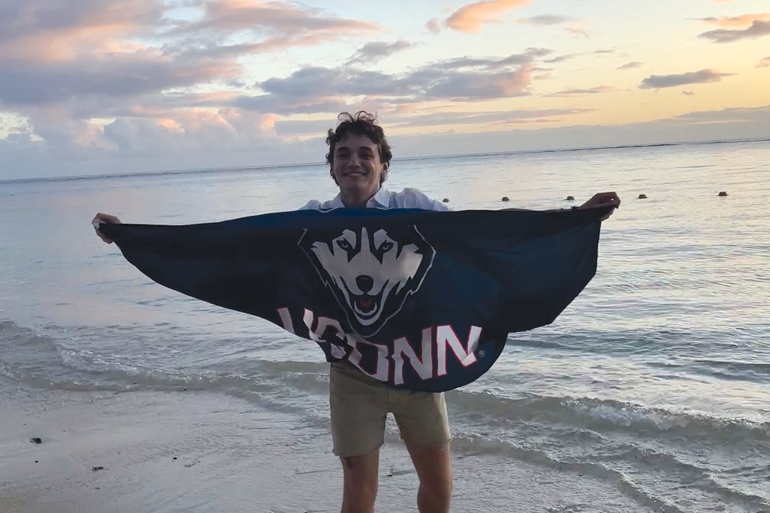 Joshua Ellenberg holds a UConn banner while his feet are in the Indian Ocean