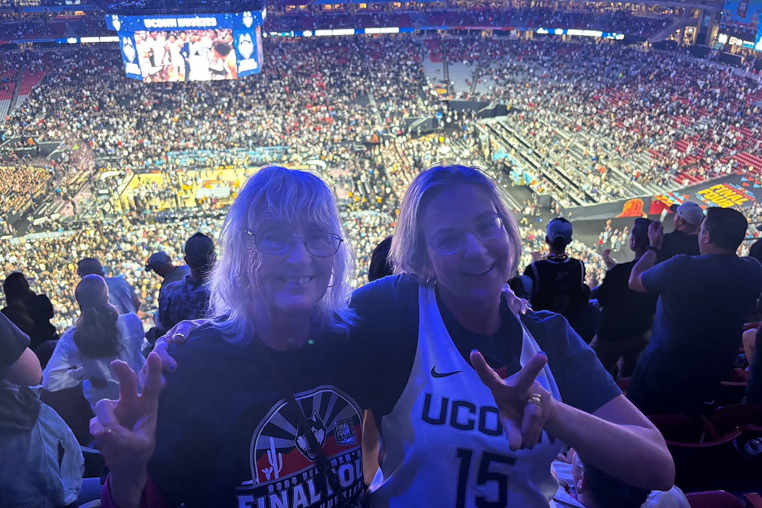 Vanessa and her mother pose with peace sign hand gestures in front of crowd gathered in seats for basketball game in Gampel Pavillion