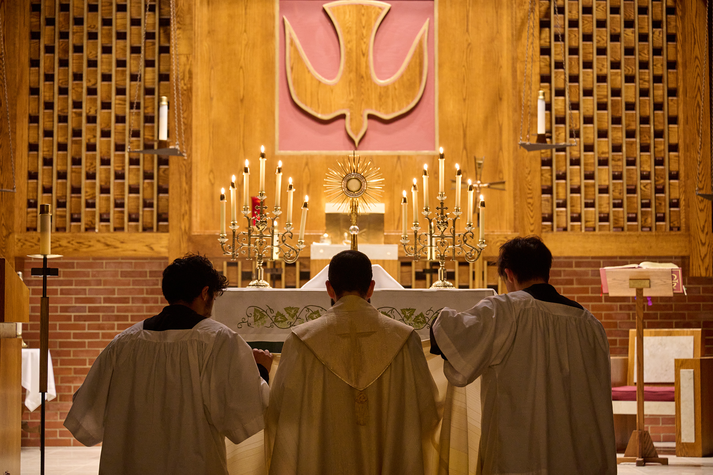 Three clergymen bow their heads in silent prayer, while candles are arranged in front, and a wooden dove shape decorates the wall.