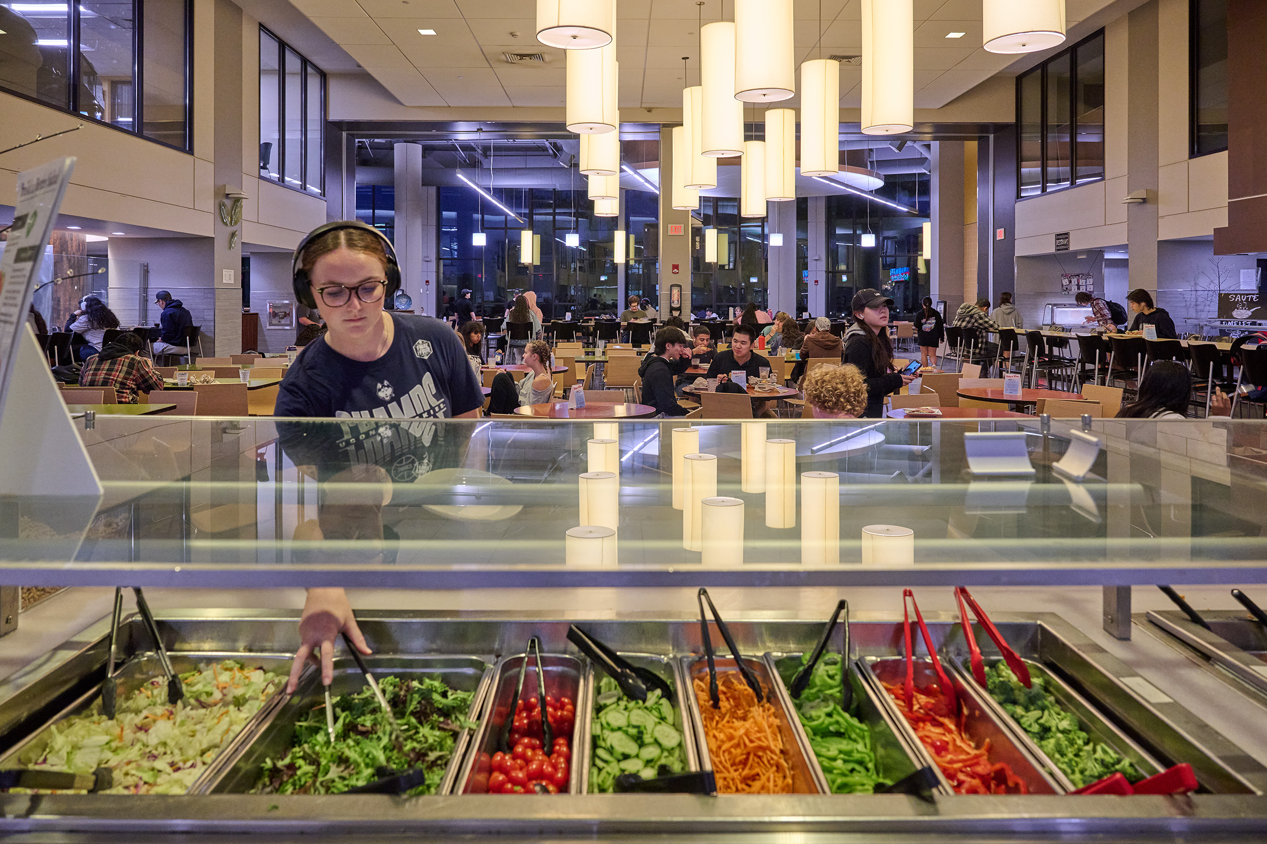 Female student wearing headphones grabs spring mix at McMahon’s salad bar, with students eating behind her.