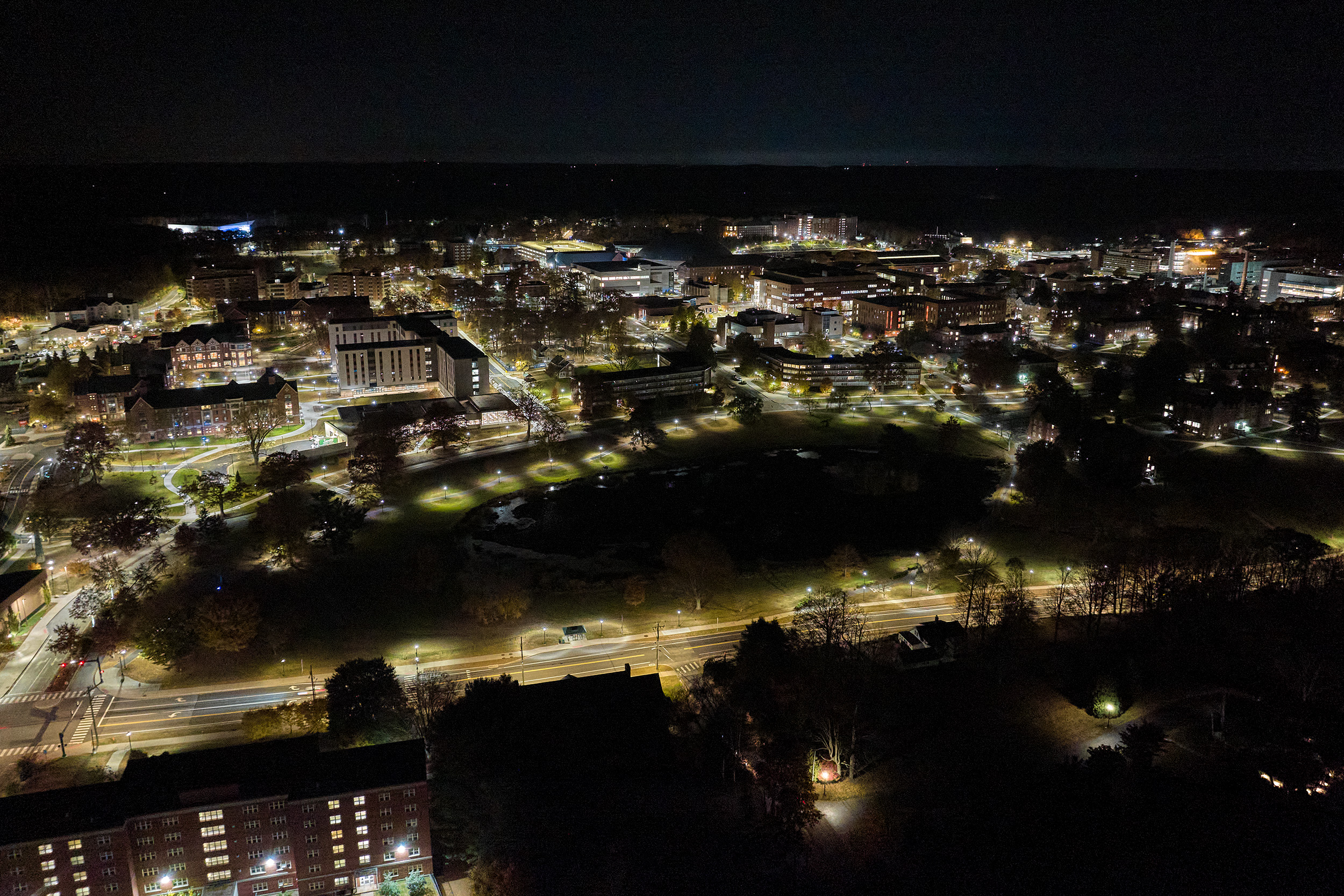 Aerial view of UConn campus at night, showing academic buildings, paths, dorms, and dining halls lit up.