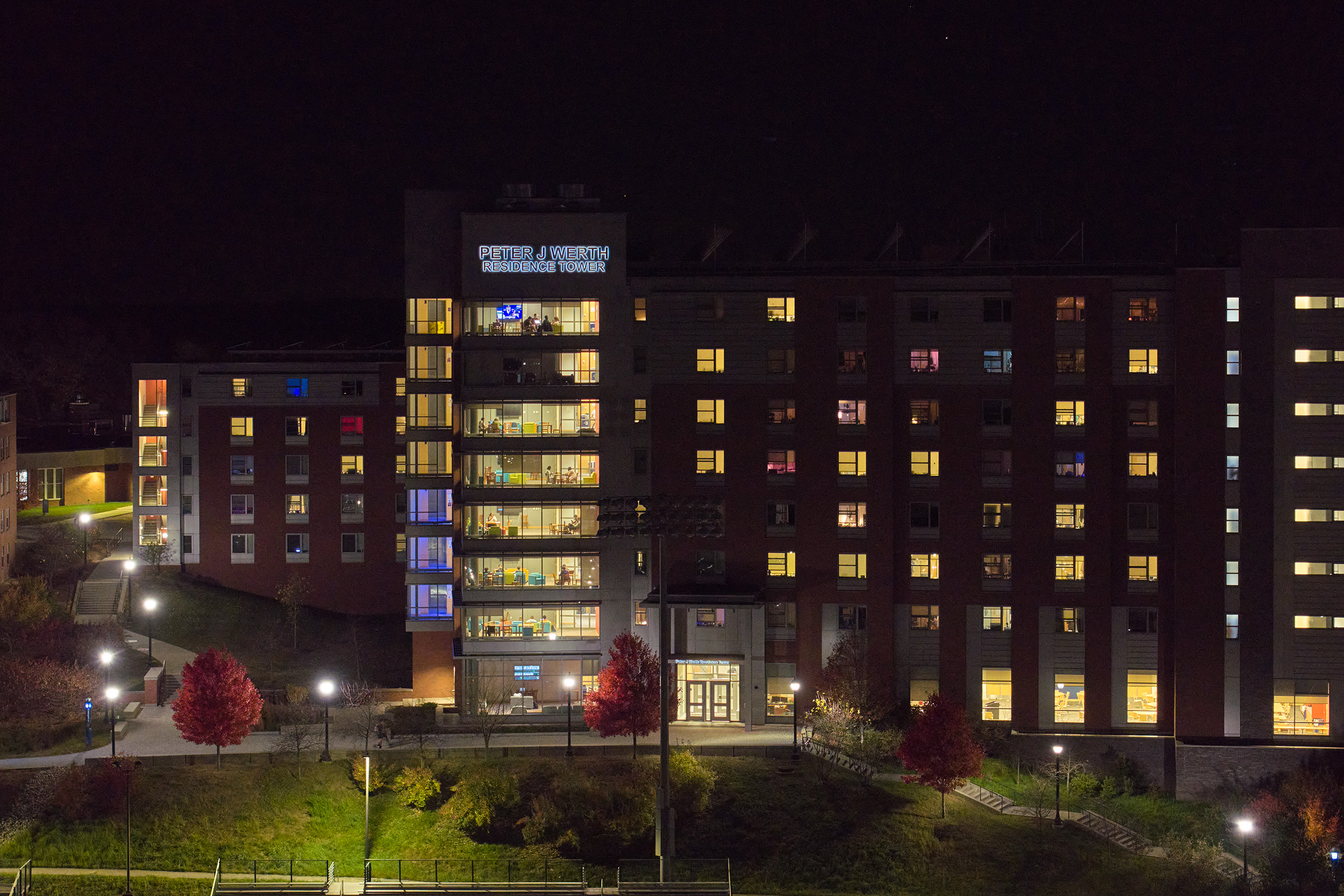 View of the Peter J. Werth Residence Tower, showing lit student lounges and dorm windows with various colored lights inside, such as blue, red, yellow, and pink.