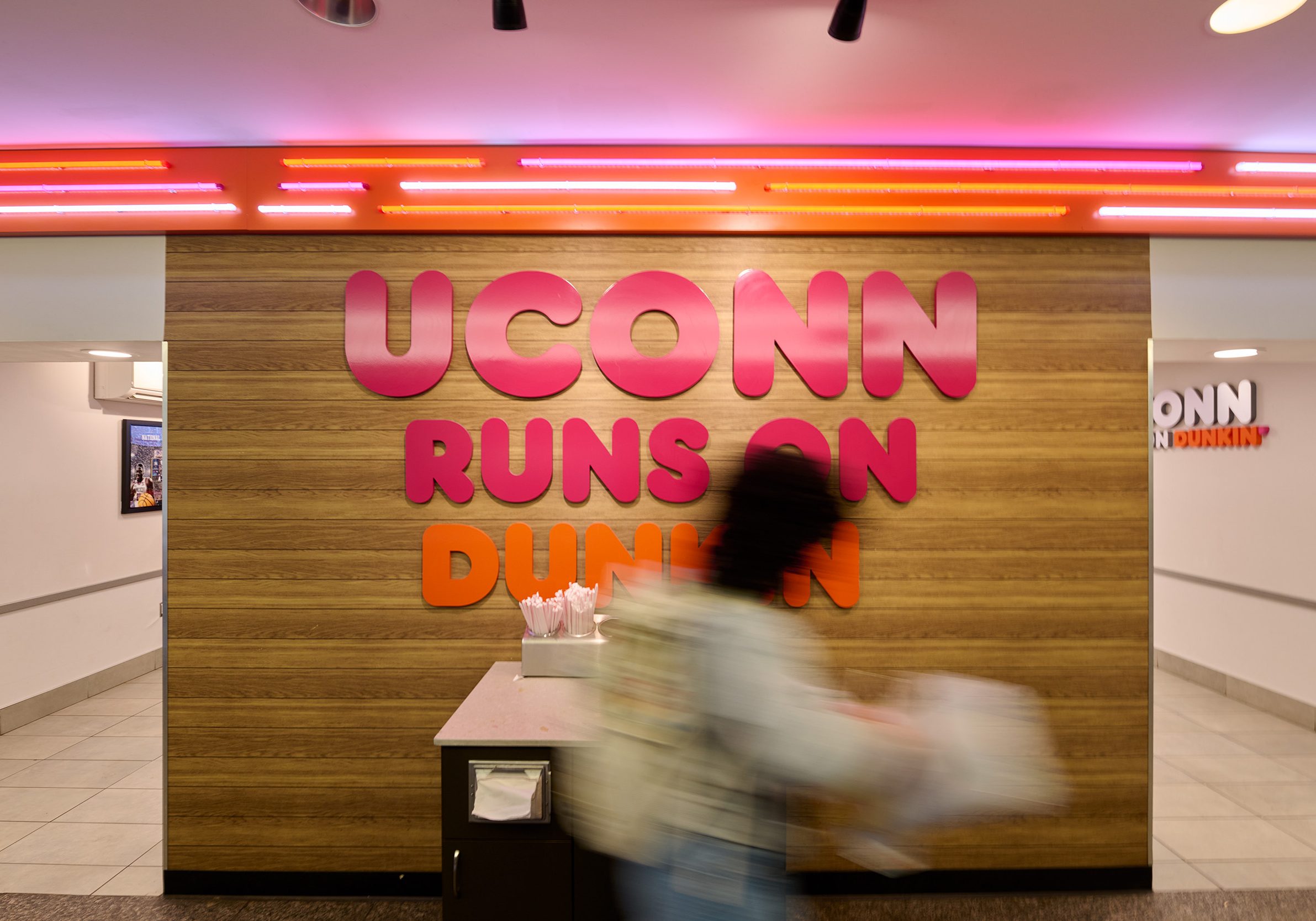 A blurry student rushes away with their order, with a vibrant pink and orange “UConn Runs on Dunkin” sign in the background at the Student Union.