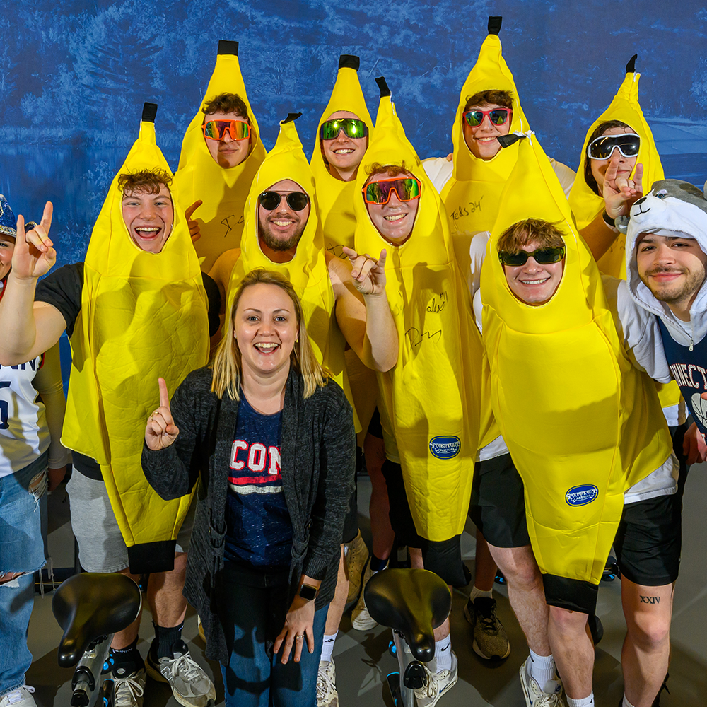 Julie Bartucca poses in front of UConn sports fans. One of the ten fans is adorned with a husky animal hat, another in a UConn blue and baddazzeled cowboy hat, and the rest (8) are dressed in banana costumes.