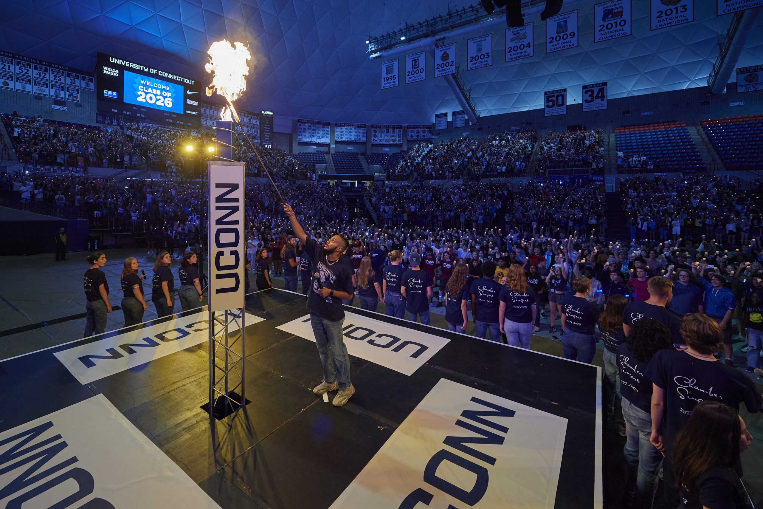 Man lighting large torch on stage. A crowd of people sitting behind him.