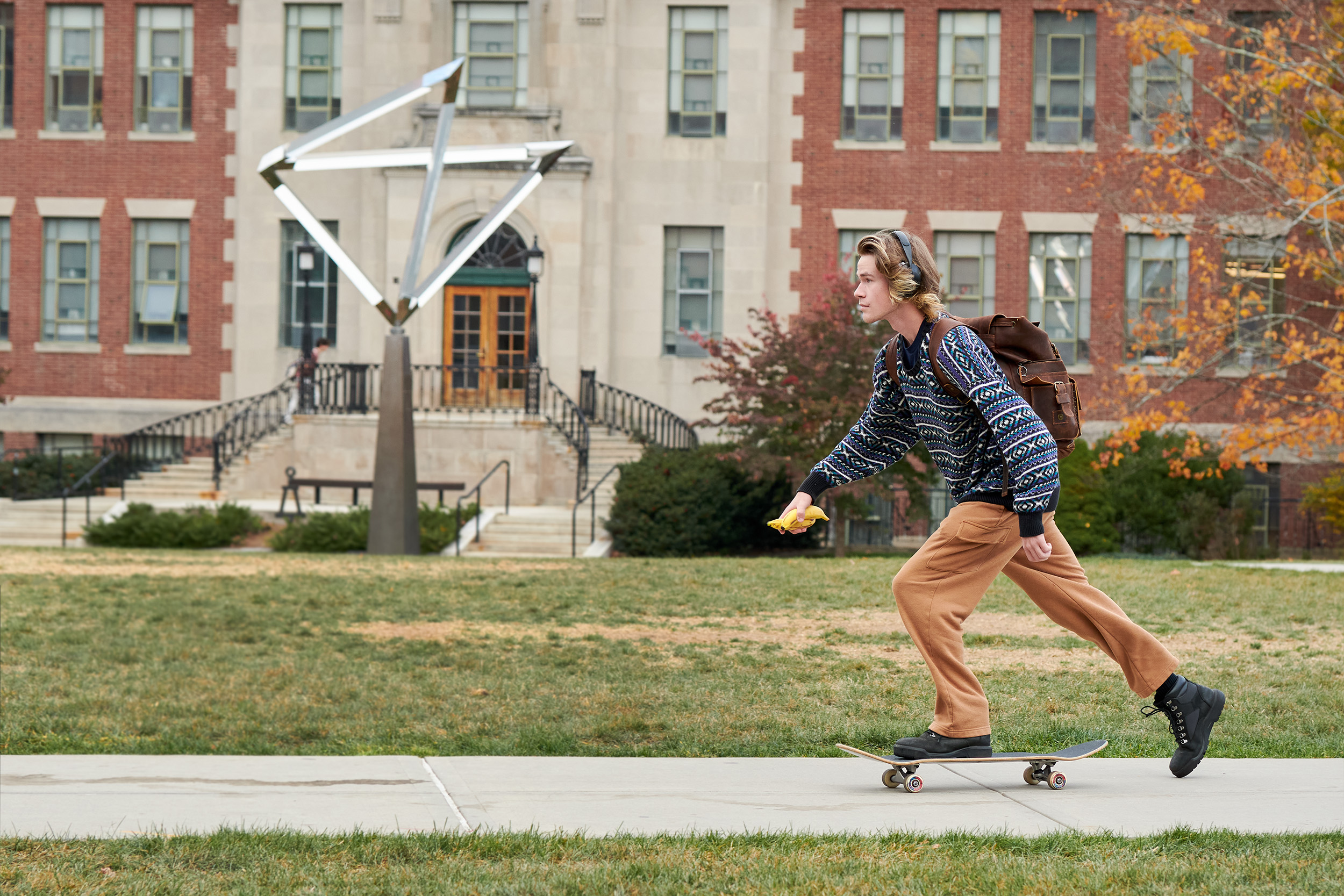 A male student wearing a patterned sweater rides a skateboard outside the Student Union while holding two bananas.