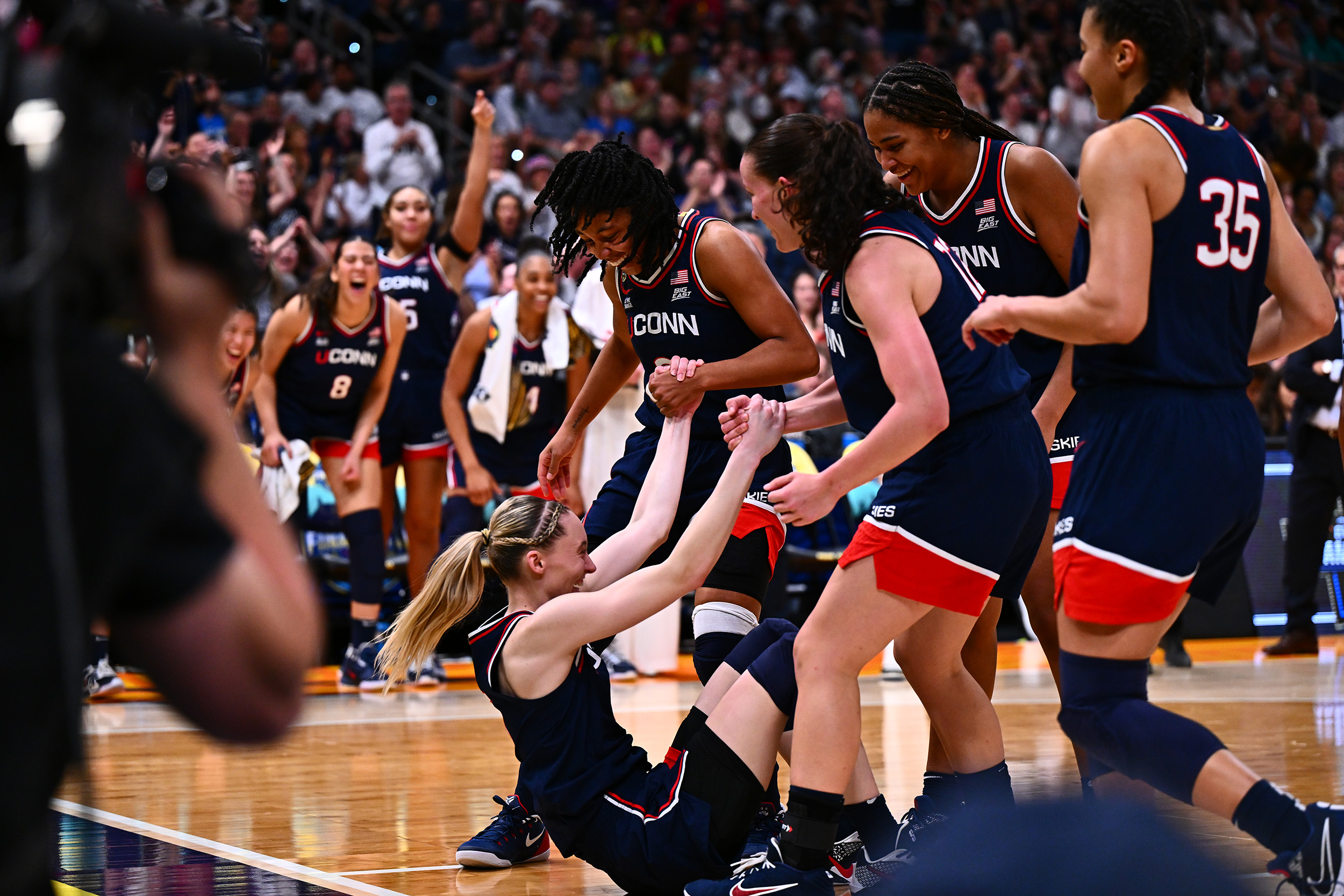 AMB_3948 A smiling Paige Bueckers is helped up off the floor by her teammates while several more cheer on in encouragement from the sideline behind them.