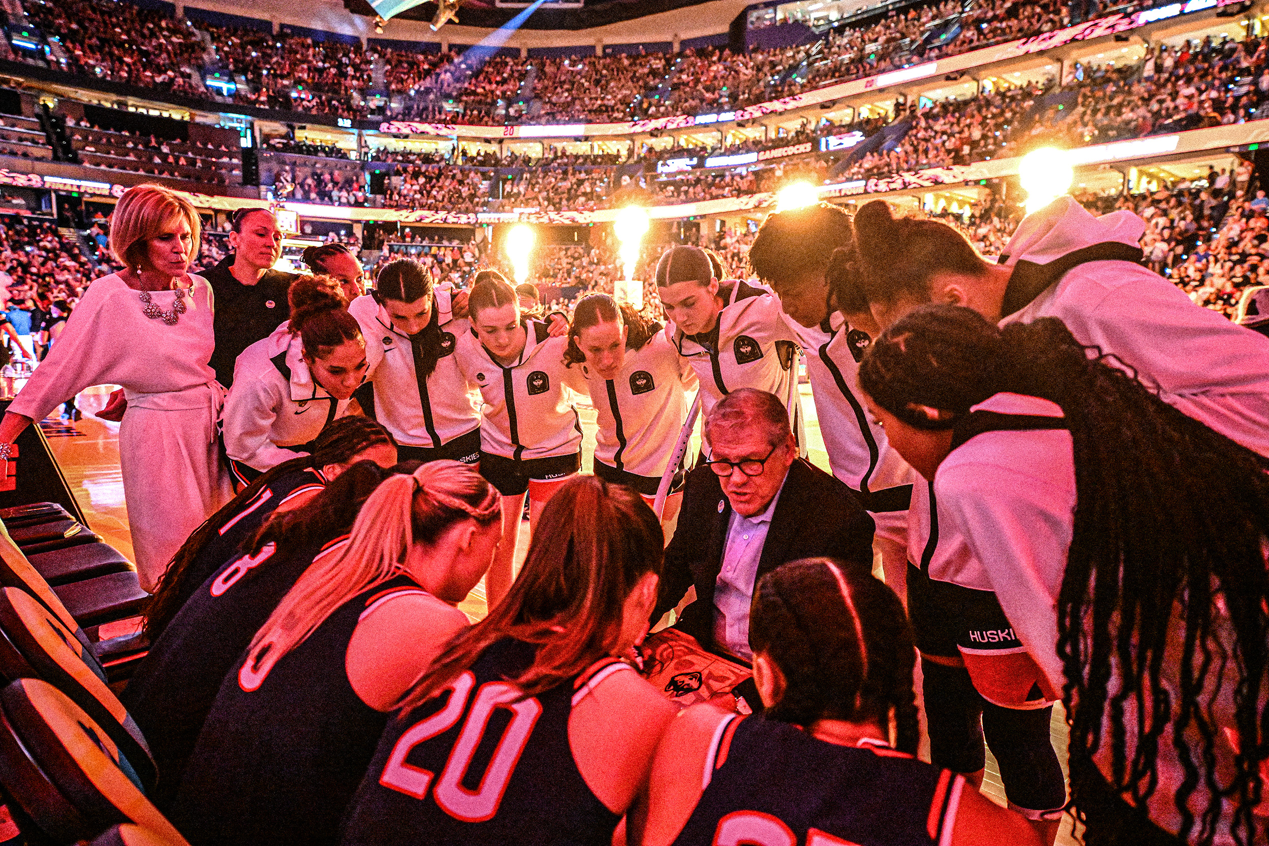 AMB_4286 The UConn women’s basketball team locks arms in a pre-game huddle surrounding head coach Geno Auriemma while pyrotechnical effects are being lit behind them in a sold-out arena.
