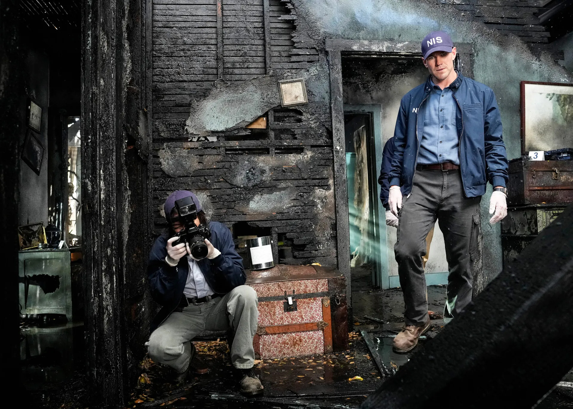 Austin-Stowell-Says-Personal-Trauma-Informed-NCIS-Origins-Performance-1_Greg Gayne_CBS Austin Stowell, right, as Leroy Jethro Gibbs during a scene from “NCIS: Origins”. Stowell’s character walks into charred building crime scene with man photographing evidence.