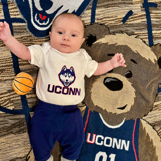 Baby Luca Robert Timko in white UConn onesie laying on top of UConn blanket with Jonathan the Husky logo and teddy bear wearing UConn jersey