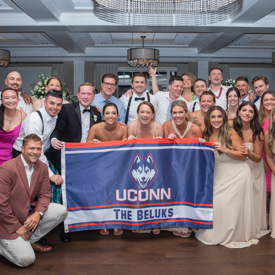 Large group of alums stand and crouch with UConn flag at Allyson (Gambino) Beluk and Michael Beluk’s wedding