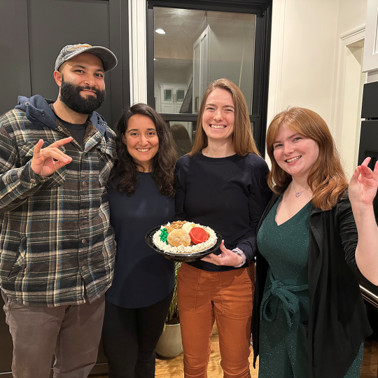 One male and three females stand together in a kitchen holding a thanksgiving pie
