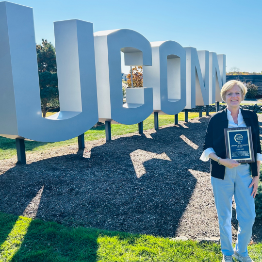 Leslie Imse holding Lifetime Achievement Award plaque in front of UConn gateway sign