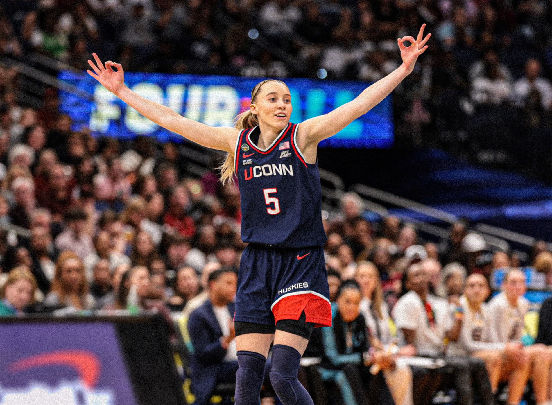 ME792045-sized-blur Paige Bueckers spreads her arms wide and holds three fingers up on each hand in celebration of a made three-point shot while South Carolina players and coaches watch from their bench behind her.