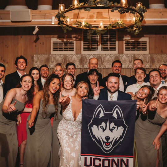 Large group of alums stand with UConn flag at Ryan Palzere and Chelsea Cocozza’s wedding