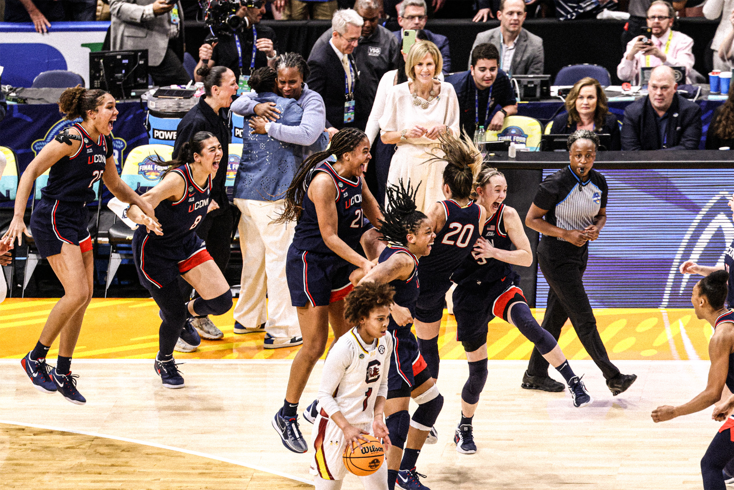 TM035256-sized UConn women’s basketball players and coaches jump into the air with excited expressions and embrace each other in celebration as the referee blows the final whistle.