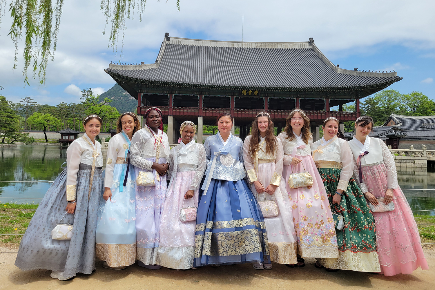 20250522_110056 A diverse group of 9 women stands in front of a Korean palace wearing Hanbok, traditional Korean dress, in blues, purples, greens, and pinks.