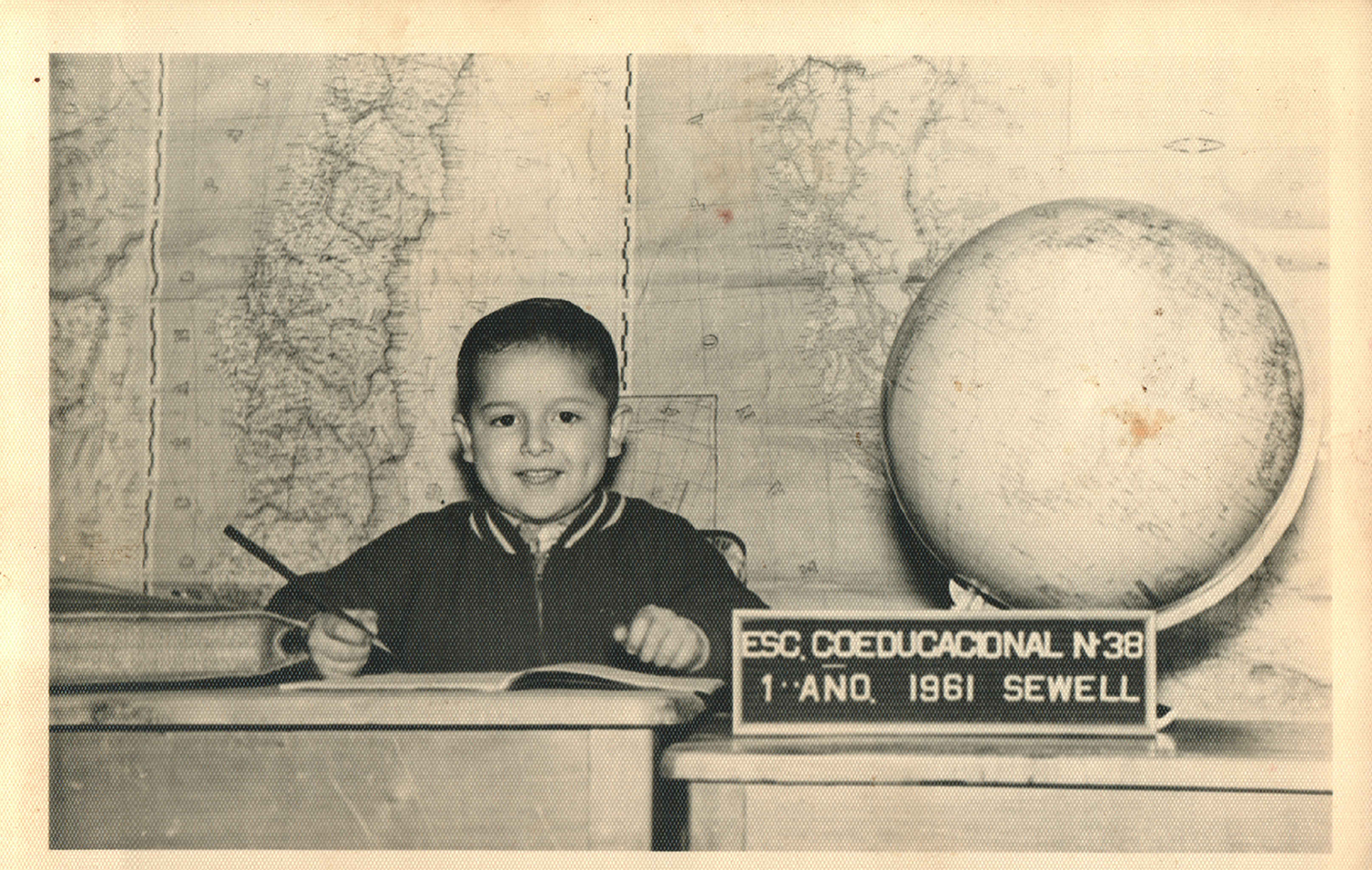 A black and white photo of a 6-year-old boy with black hair sitting at a desk, smiling at the camera and holding a pen. He is in front of a map and next to him are a globe and a sign that reads: ESC. Coeducacional N 38 1 Ano 1961 Sewell