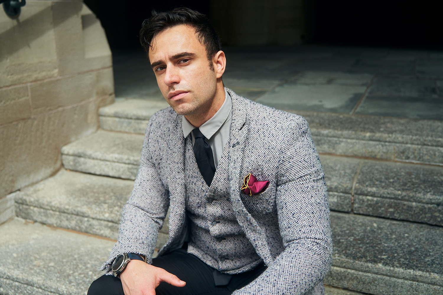 Dieter Tejada sits on stone steps wearing a tweed suit with a maroon pocket handkerchief and dark tie, looking pensively at the camera