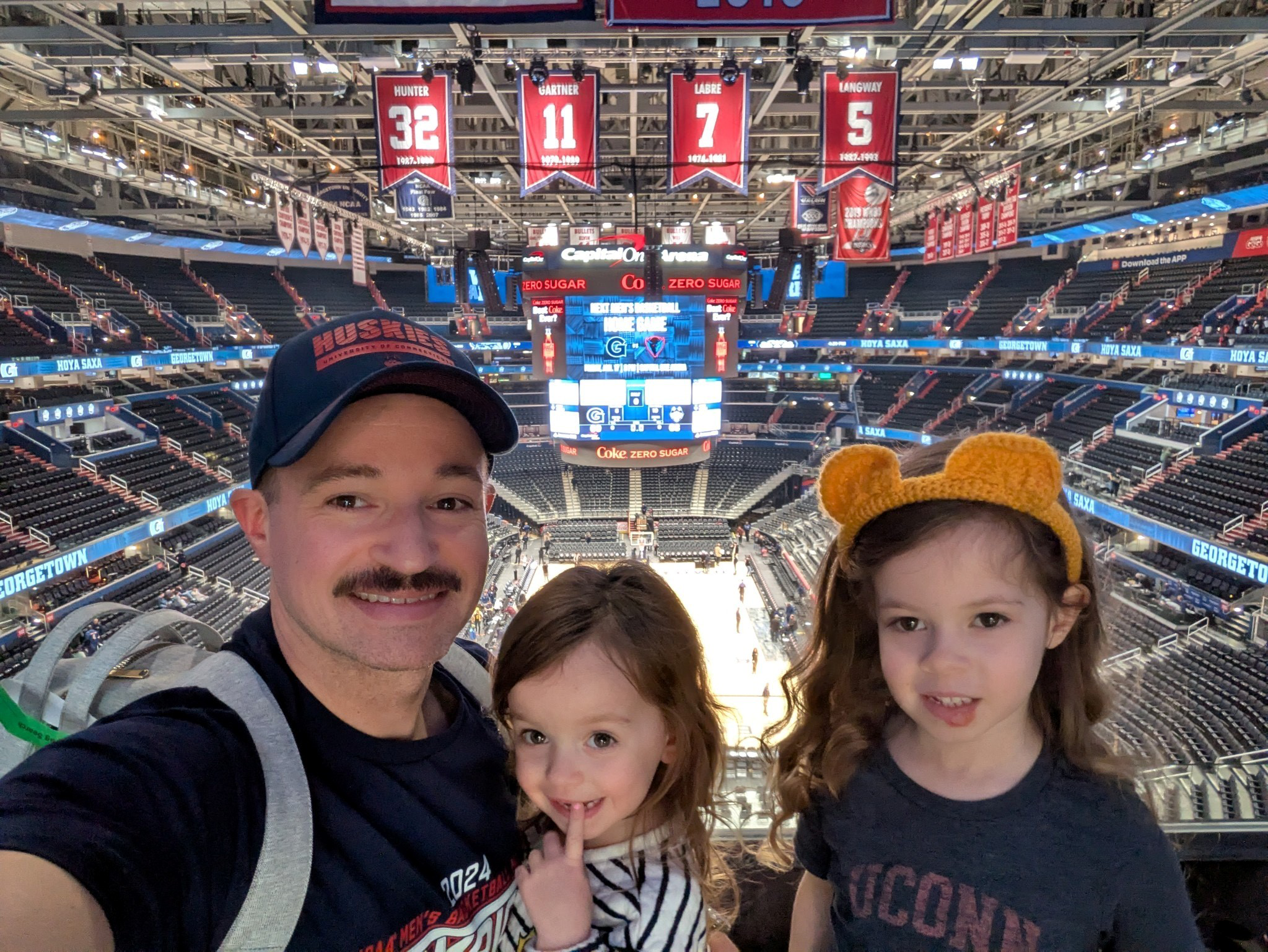 George Philbrick, wearing a Huskies hat and T-shirt, with his two young daughters in a mostly empty Capital One arena with red flags hanging from the rafters.