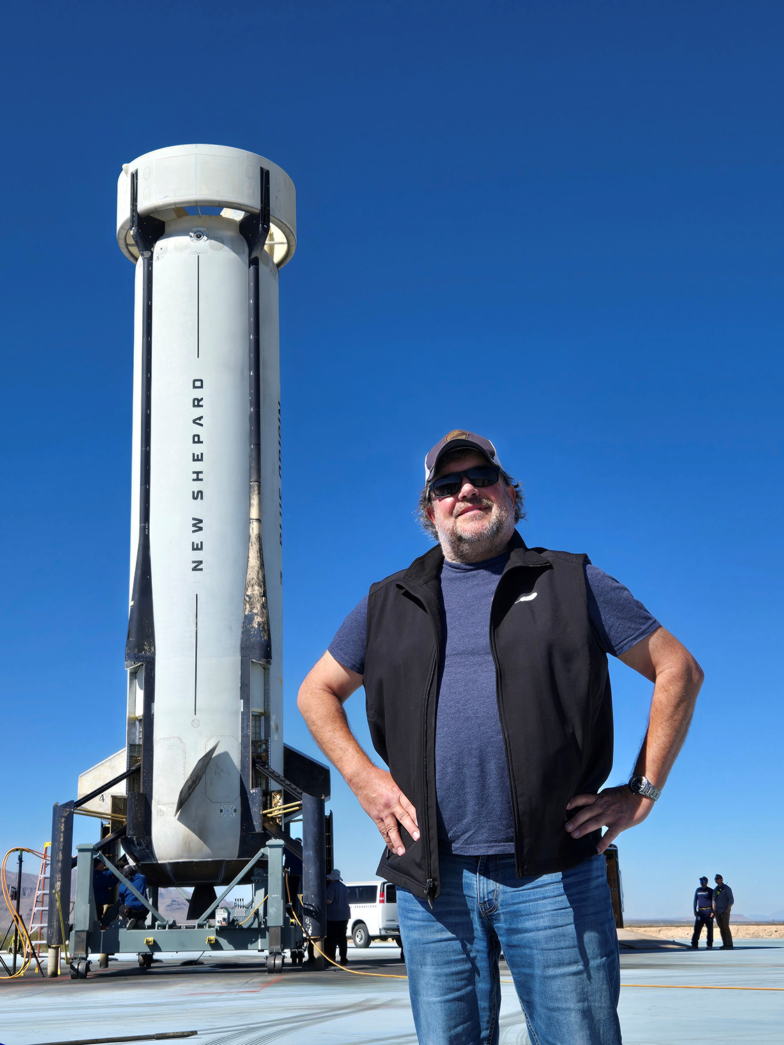 David Smail poses with his hands on his hips in front of a blue sky and a Blue Origin rocket with “New Shepard” written on the side 