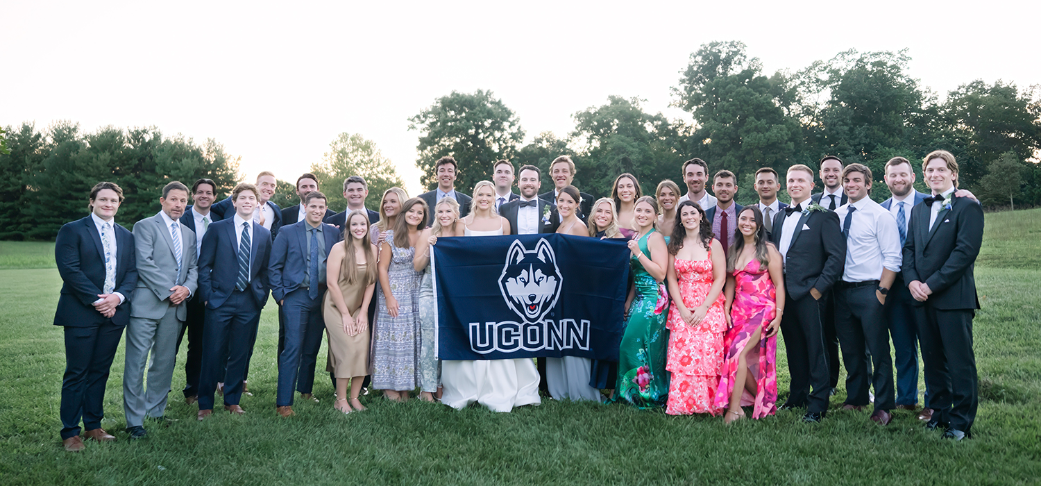 737467763-hayden-atwater-wedding – A large group of men and women in wedding attire pose in a field holding a UConn banner 
