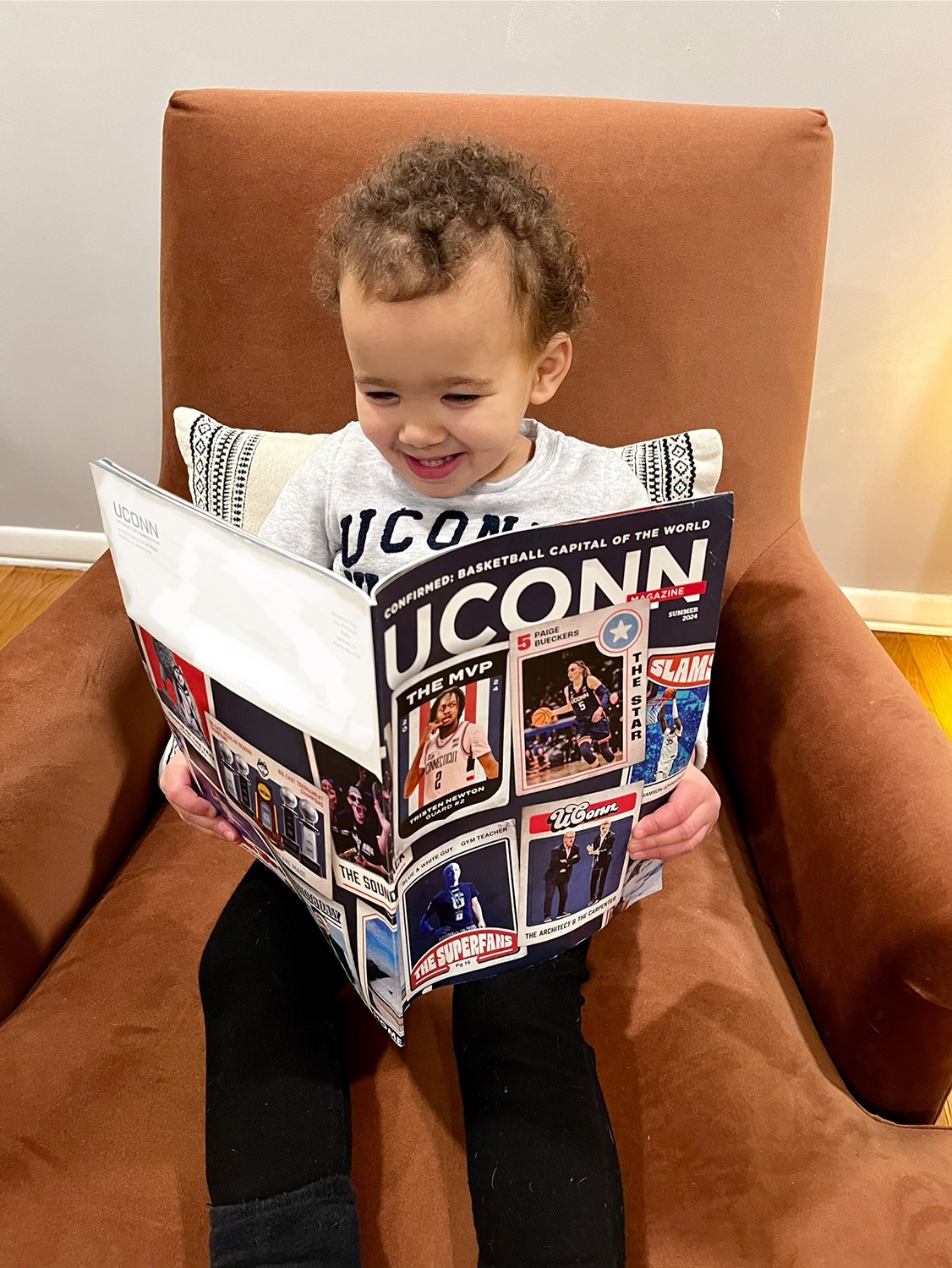 737467782-kaitlyn-herman-jessee-s-baby-colbie-jessee Colbie Jessee, 2, smiles in an armchair as she looks at UConn magazine with basketball cards on the cover