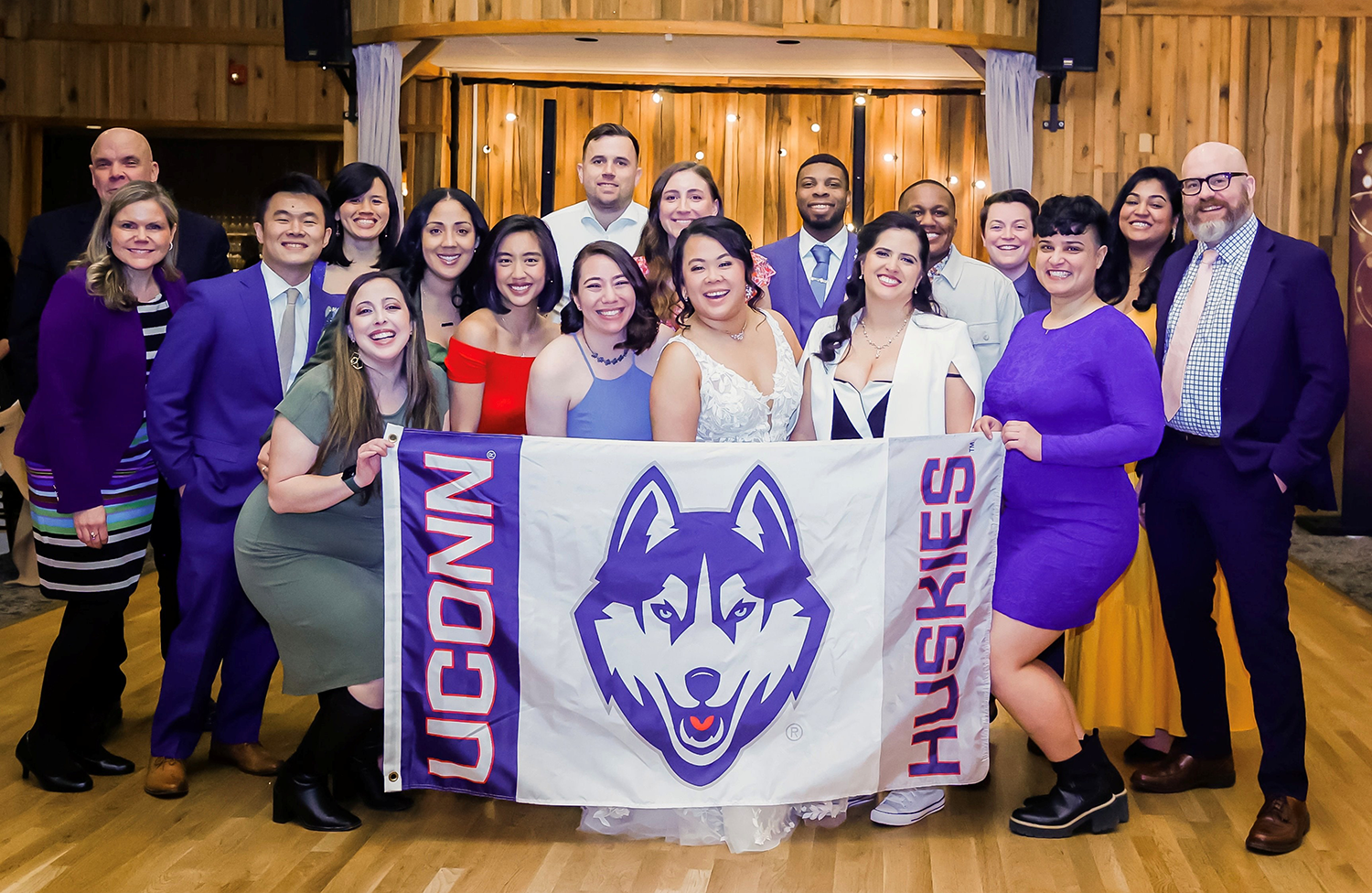 A group of men and women mostly wearing purple and white pose with a UConn Huskies banner on the dance floor at a wedding