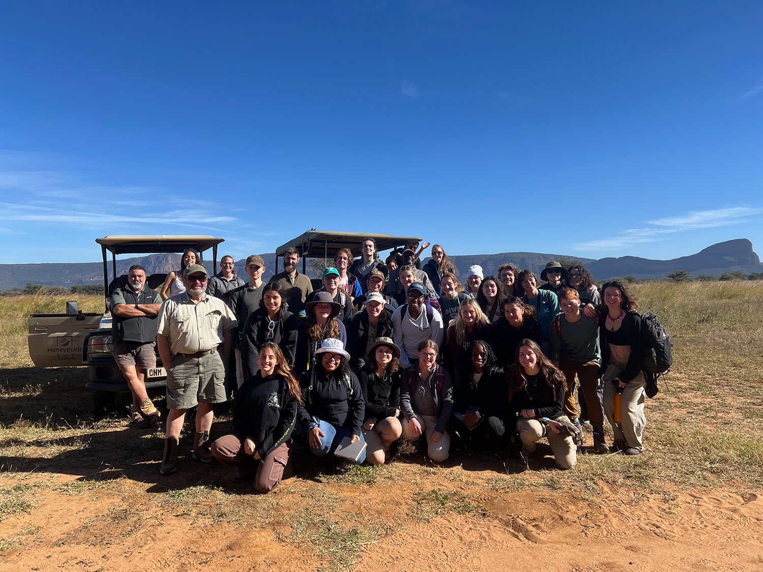 A large group of students with Morty Ortega and local guides pose in front of two open-air jeeps in a grassy field with mountains in the background and a blue sky
