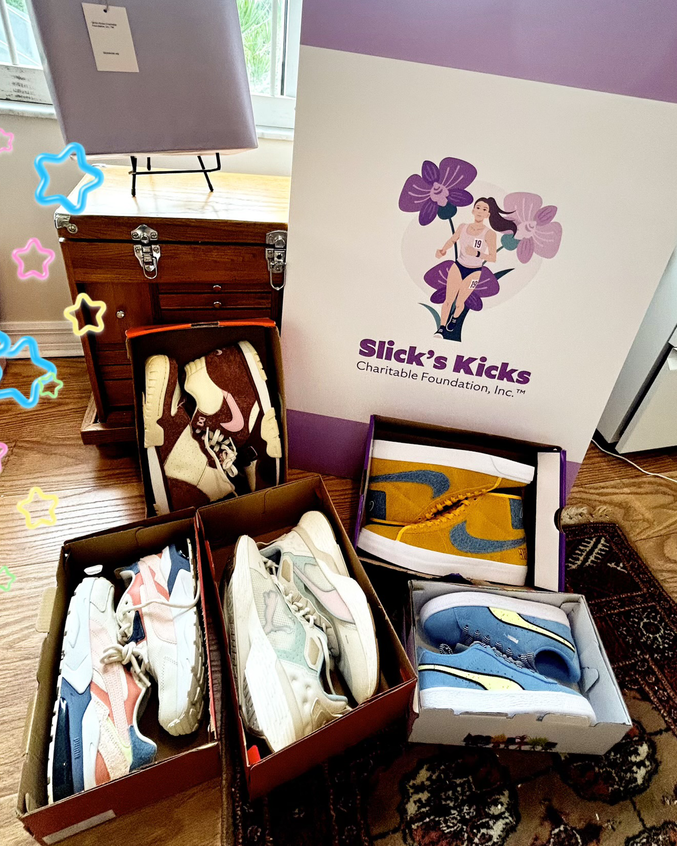 B8A89FB5-1918-43BF-B5B4-FF4011F63C3C 5 different colored pairs of sneakers each in their own box with a poster behind them that says “Slick’s Kicks Charitable Foundation, Inc.” with an illustrated logo of a woman running with purple flowers behind her
