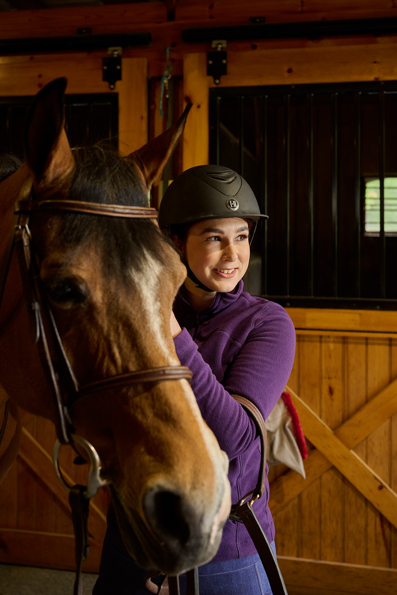 In a stable, Brooke Gemmell, wearing a black riding helmet and purple top, smiles and looks slightly off camera next to a brown horse.