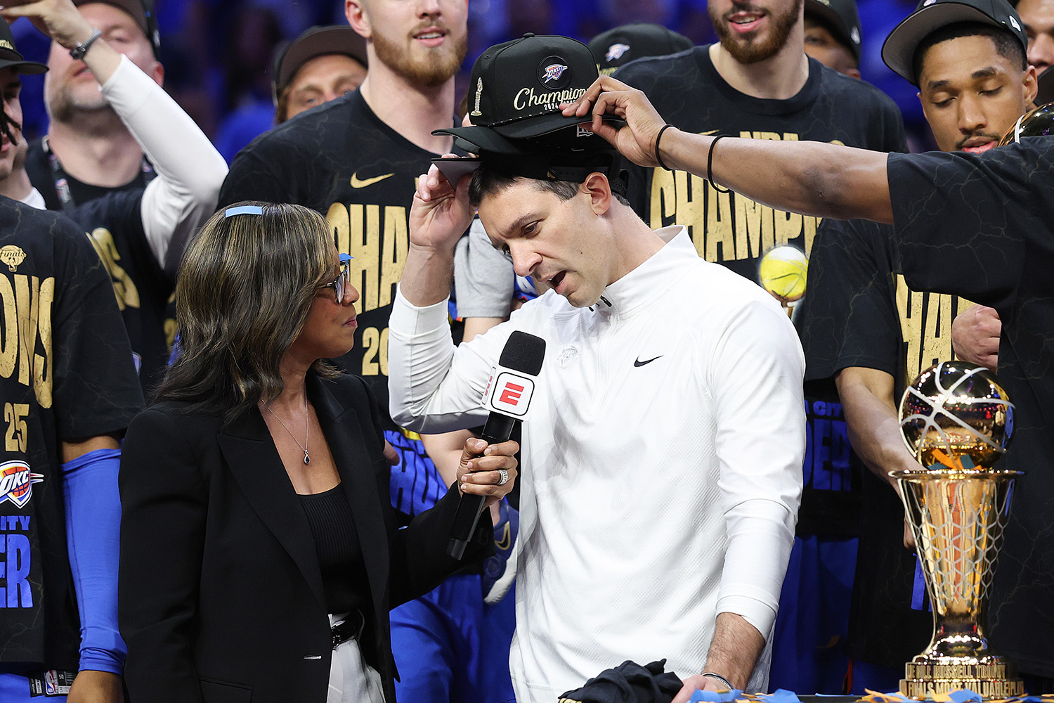 Mark Daigneault talks to an ESPN reporter with the NBA Championship trophy to his left and a player putting a Champion hat on his head