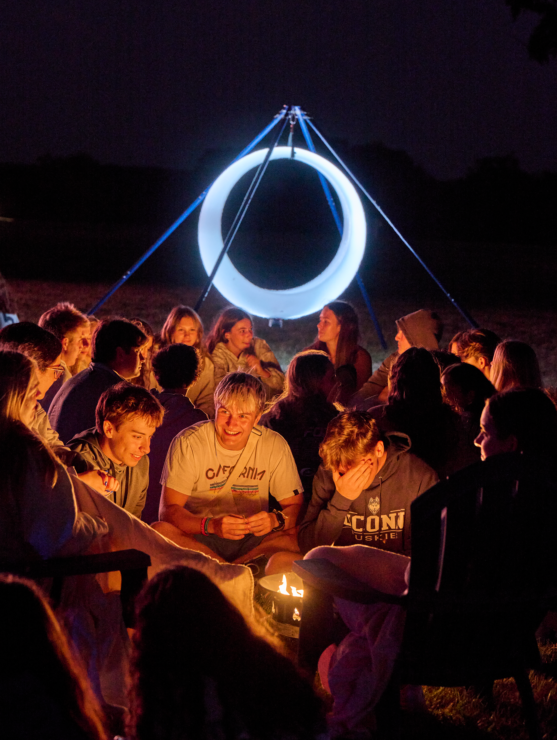 In the foreground, three male students are smiling while talking with others around a fire pit, another group sits around another fire pit behind them. A glowing O-shaped swing stands behind them against a night sky