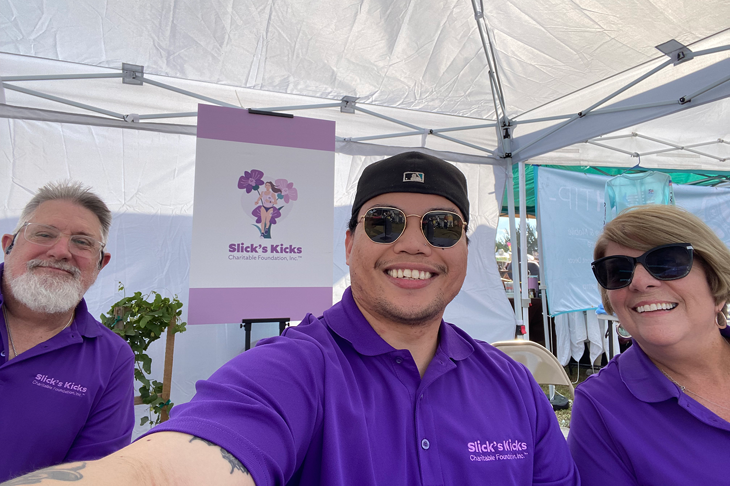 IMG_1493 Two men and a woman wearing purple “Slick’s Kicks Charitable Foundation Inc.” polo shirts smile while taking a selfie in front of a Slick’s Kicks poster