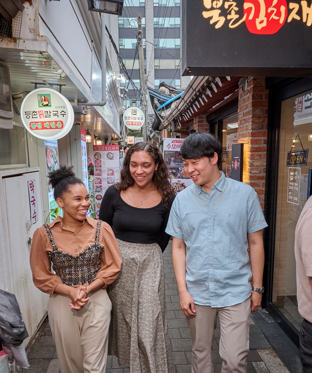 Korea_240622a012 Nadejah Green, Chelsea Hopson, and Mike Nelson smile and talk while walking down a Korean street with Korean words on the building signs around them