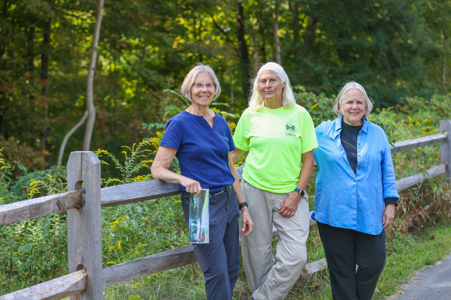  Master gardeners Karen Berger, Holly Hambleton, and Michele Jenks pose along a wooden fence with plants and trees in the background
