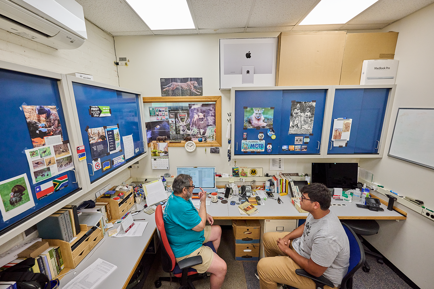 Professor Morty Ortega talks to a male student in his office, which has blue cabinets dotted with pictures of animals, different country’s flags, and other ephemera