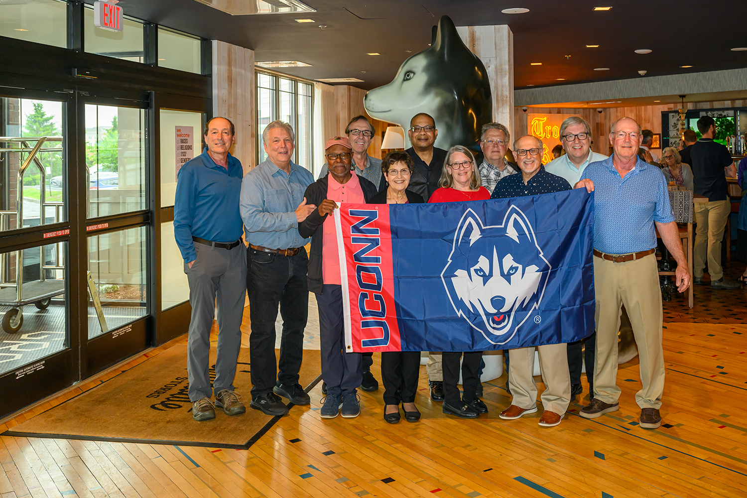 Howard Eisenberg, Dave Olsen, Martin Adomat, Richard Lowe, Carl F. Bard, Jack Havelick, Theodore R. Crockett Jr., Gail Havelick, Priscilla Drake Bard, Gary Mangiulli, and Bill McGrath pose with a UConn banner in front of a large husky dog statue in the lobby of The Graduate Hotel in Storrs