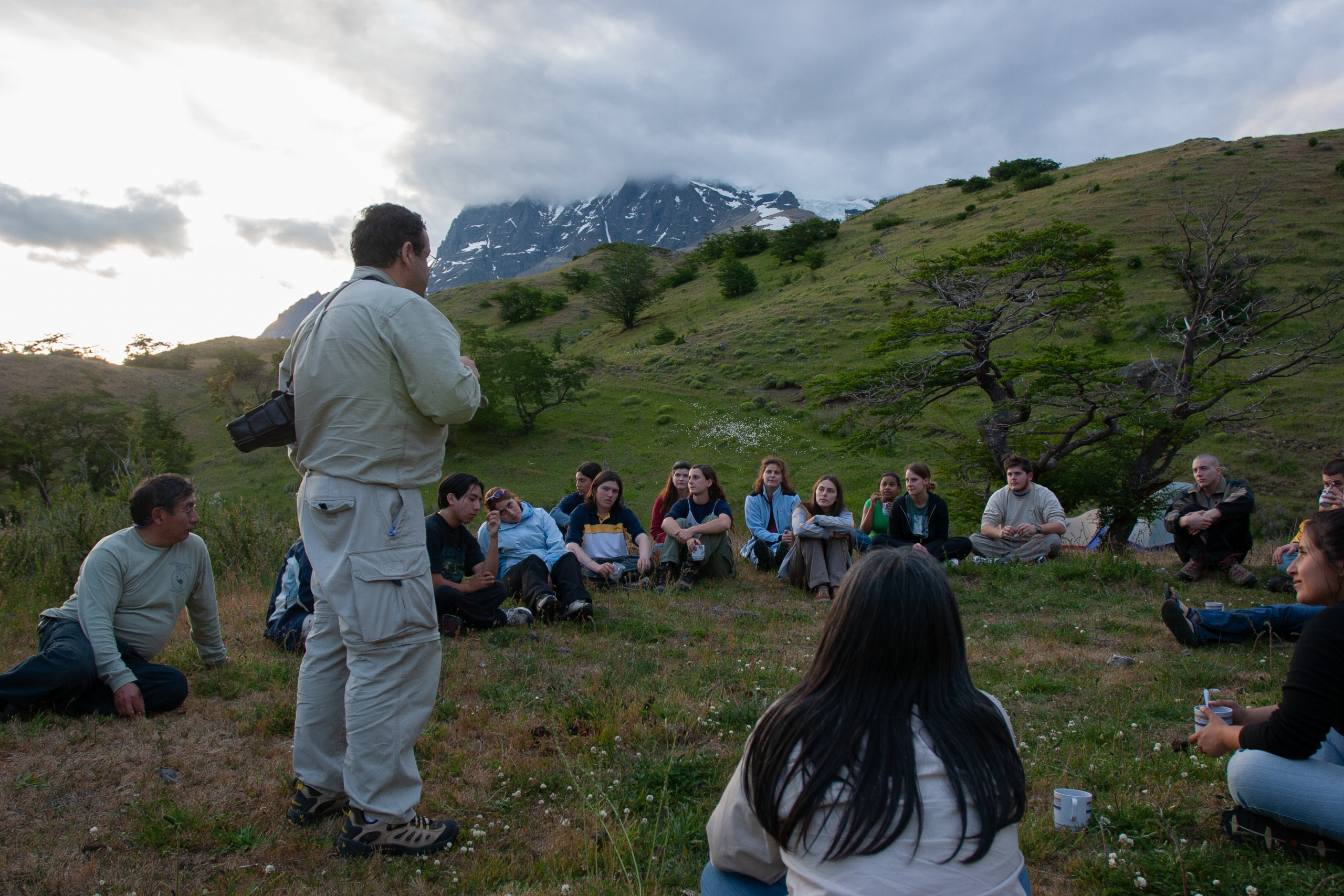 Morty Ortega, with a camera over his shoulder wearing hiking gear, stands facing a students who are sitting in a circle formation on a trip to Patagonia in 2005