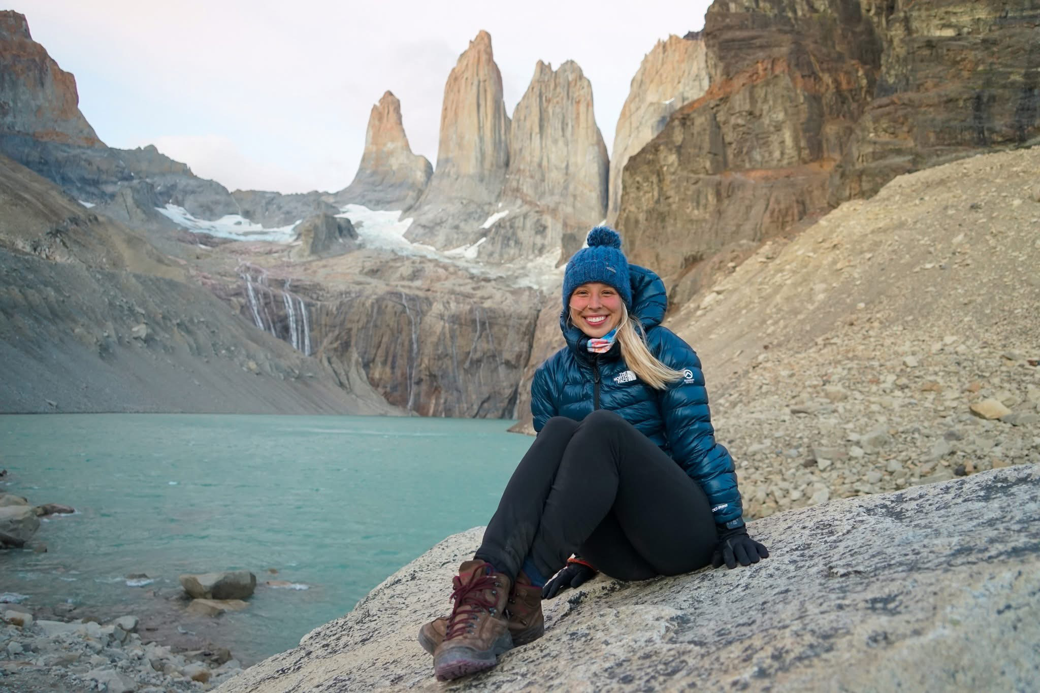 Summer Hoogenboom wearing a blue winter hat, blue North Face puffy coat, black pants, and brown boots, smiling and sitting on a rock in front of water and mountains at Torres del Paine National Park in Patagonia, Chile