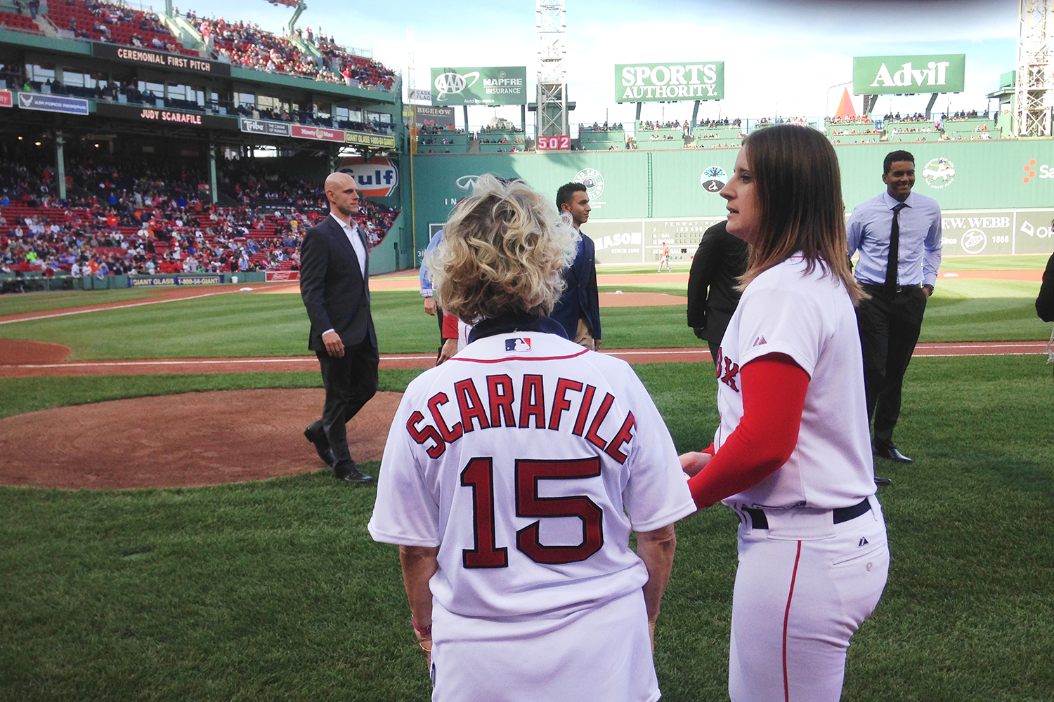 Judy Scarafile with her back to the camera, wearing a Scarafile 15 Red Sox baseball jersey, walks onto the field at Fenway Park with a woman wearing a Red Sox uniform and men in suits in the background