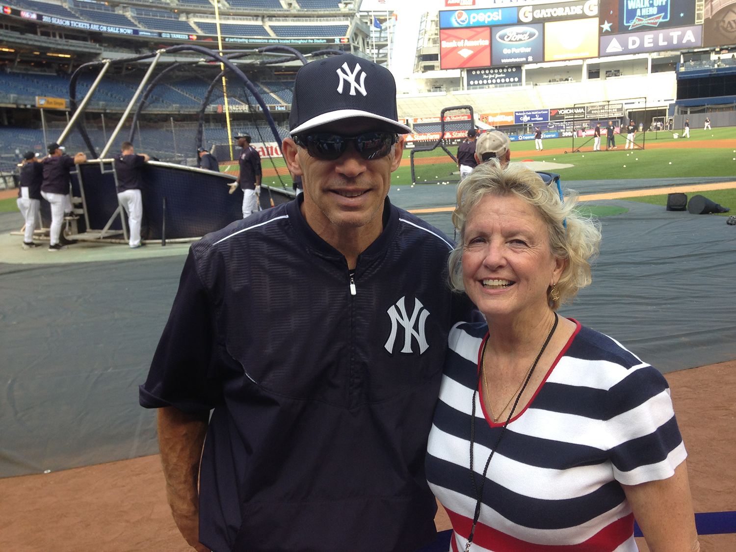 Judy Scarafile smiles with former MLB player and manager Joe Girardi on the field at Yankee Stadium with players warming up in the background